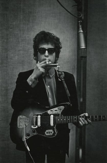 Bob Dylan in the studio with electric guitar, harmonica rack, and cigarette — black and white.